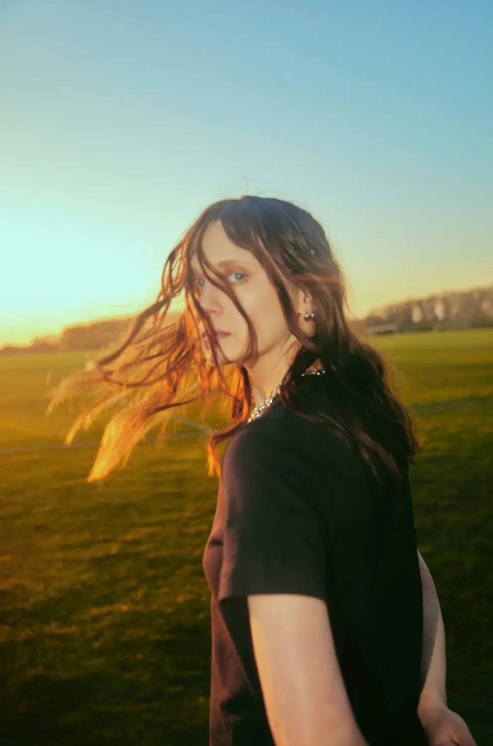 Portrait of artist Scout at sunset with windblown hair in a field, wearing a black shirt and silver jewelry